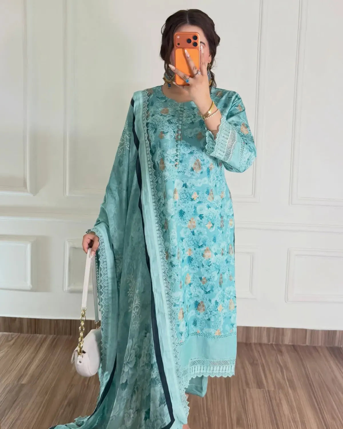 A woman takes a mirror selfie wearing a light blue-green embroidered kurta set with a matching dupatta. The outfit features delicate floral embroidery in gold and beige, with lace trim on the sleeves and hem.