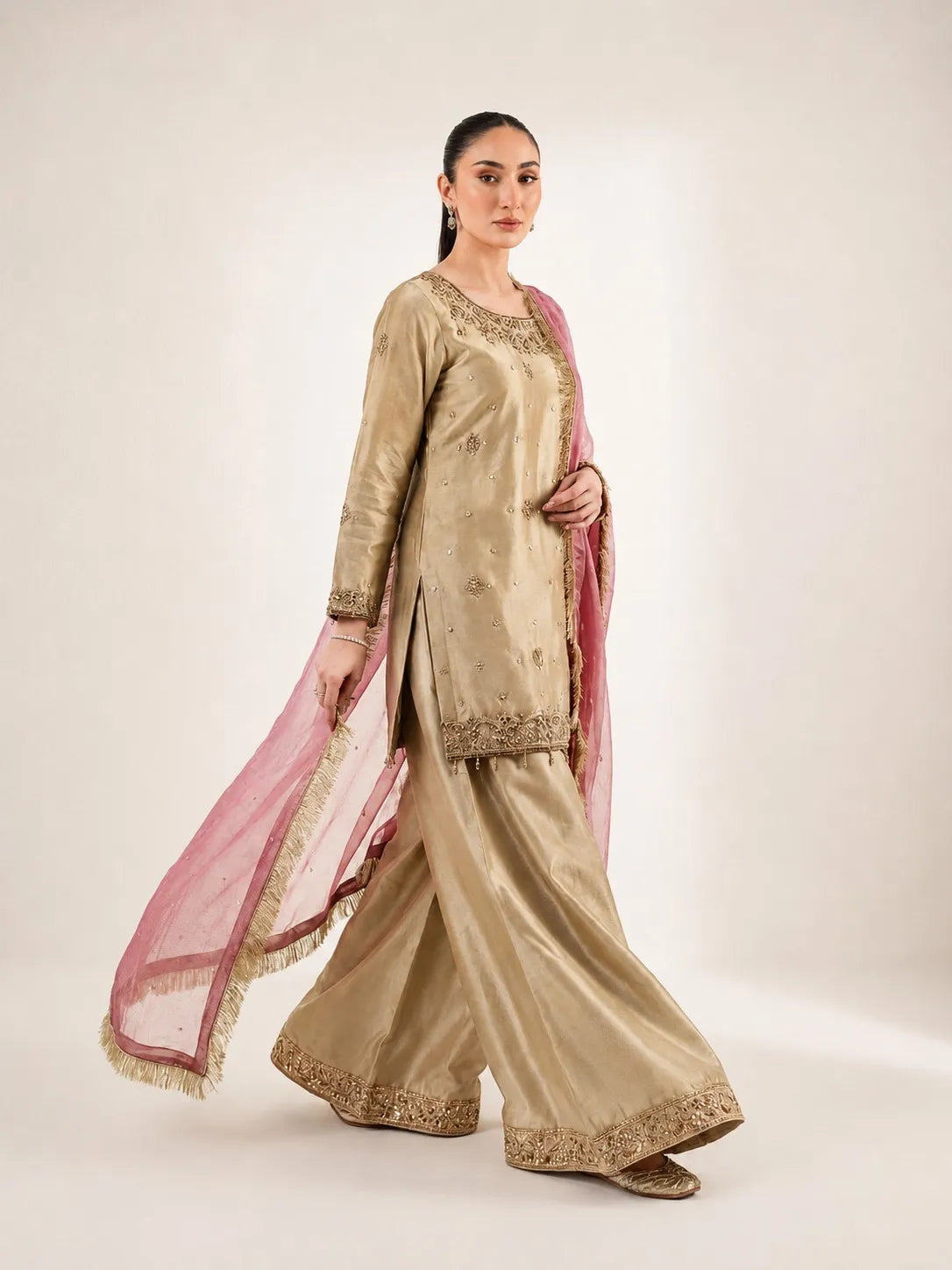 A woman modeling a traditional beige shalwar kameez suit with gold embroidery and a pink dupatta, photographed against a neutral studio background.