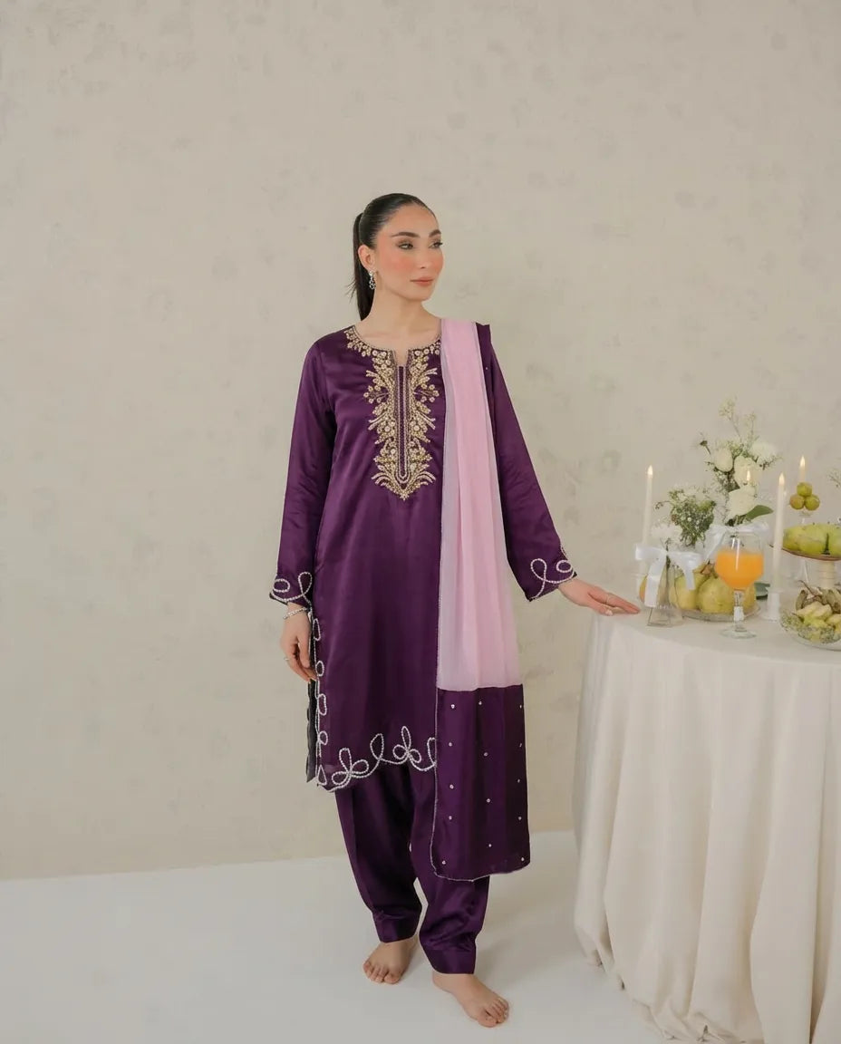 A woman modeling a traditional purple salwar kameez suit with gold embroidery, a light pink dupatta, and white embroidered details, standing next to a decorated table.