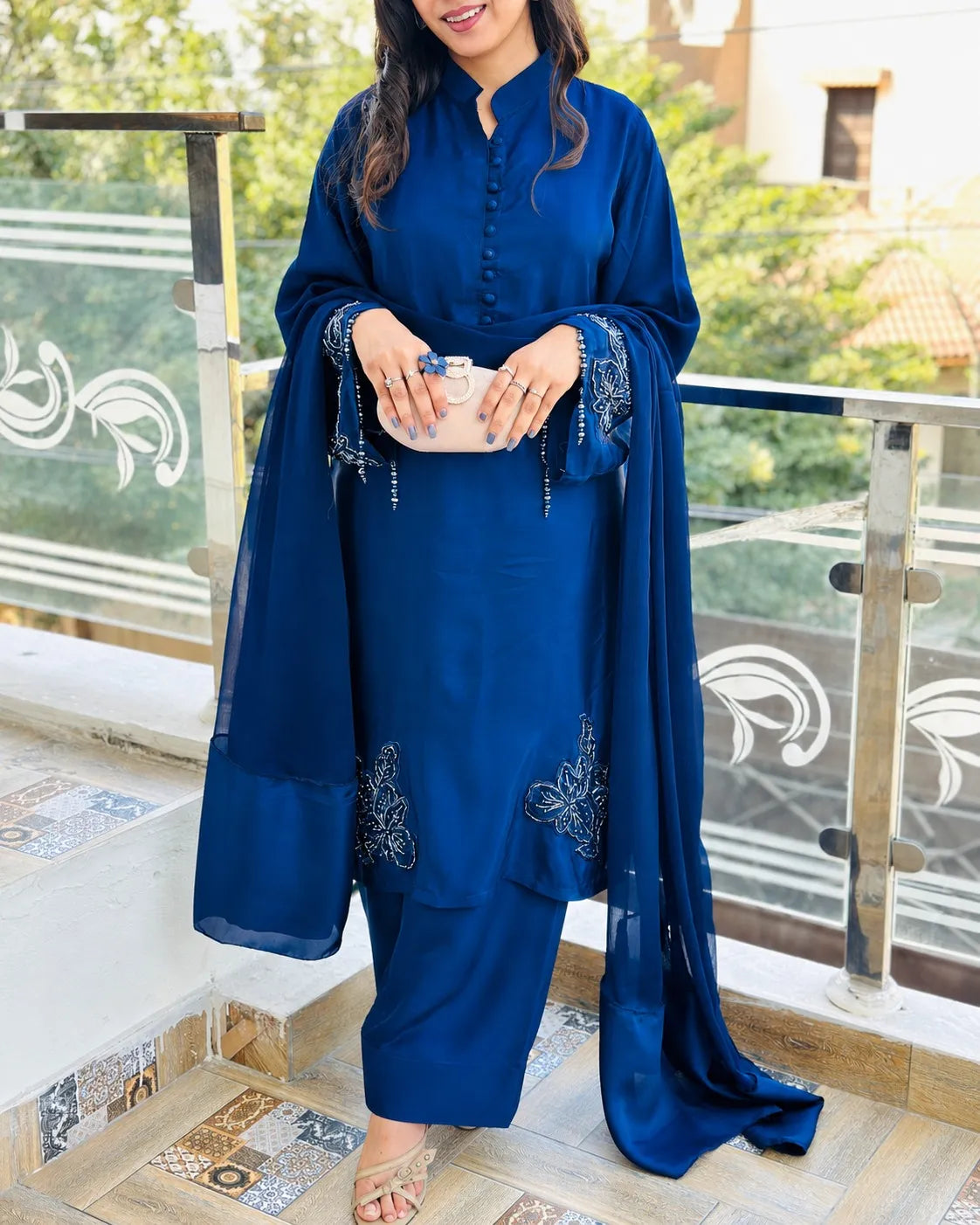 A woman wearing a royal blue three-piece traditional outfit with a matching dupatta, standing on an outdoor balcony. The outfit features a buttoned mandarin collar top, matching pants, and embroidered details on the sleeves and lower skirt.
