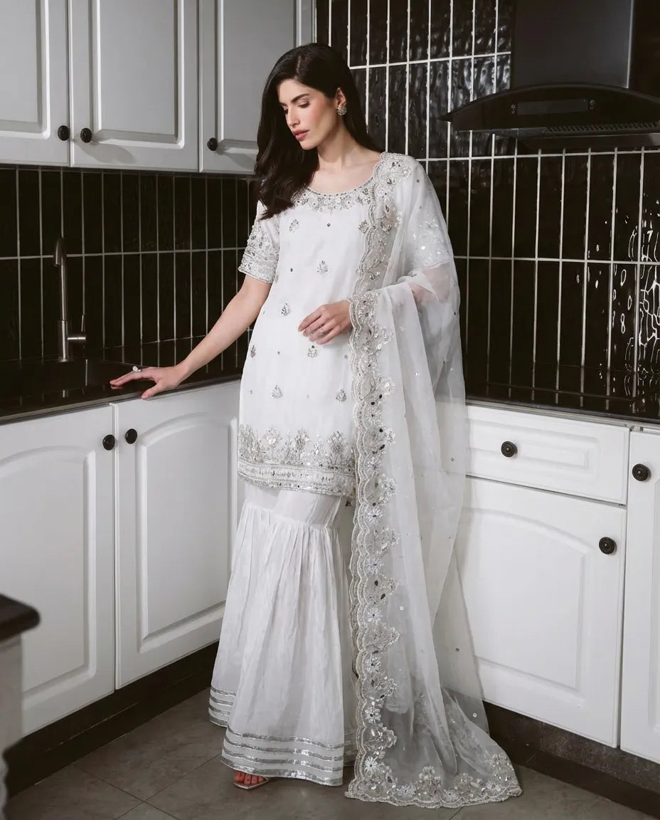 A woman wearing a white embroidered traditional outfit with a sheer dupatta stands in a kitchen with white cabinets and black tiled backsplash.