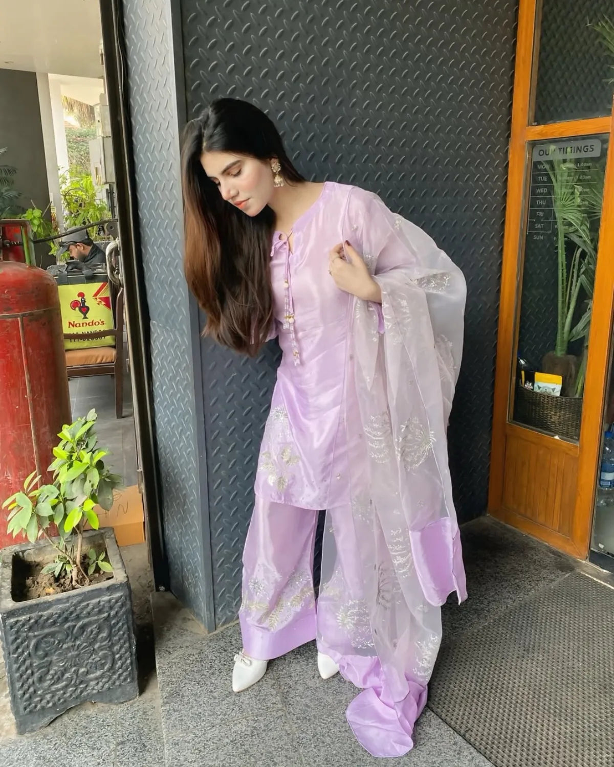 A woman wearing a light purple/lavender traditional outfit with a matching sheer dupatta, standing in front of a dark gray textured wall.