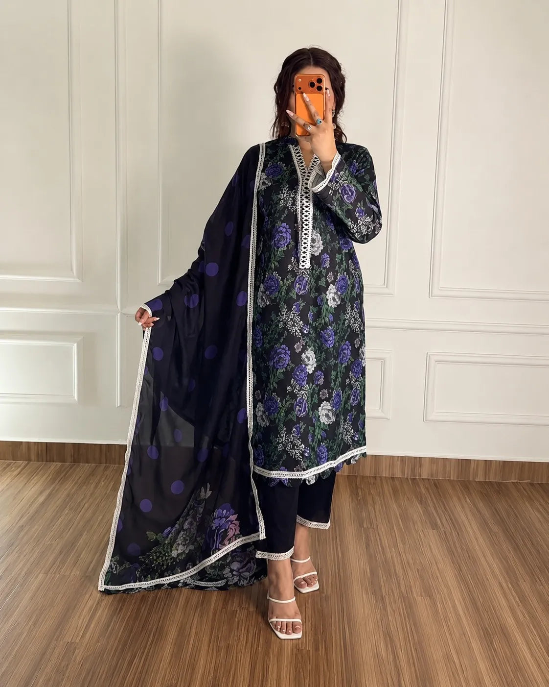 A woman modeling a black floral salwar kameez suit with a matching dupatta. The suit features purple and white floral patterns, white embroidered trim along the neckline and hem, and a black dupatta with purple polka dots and white border trim.