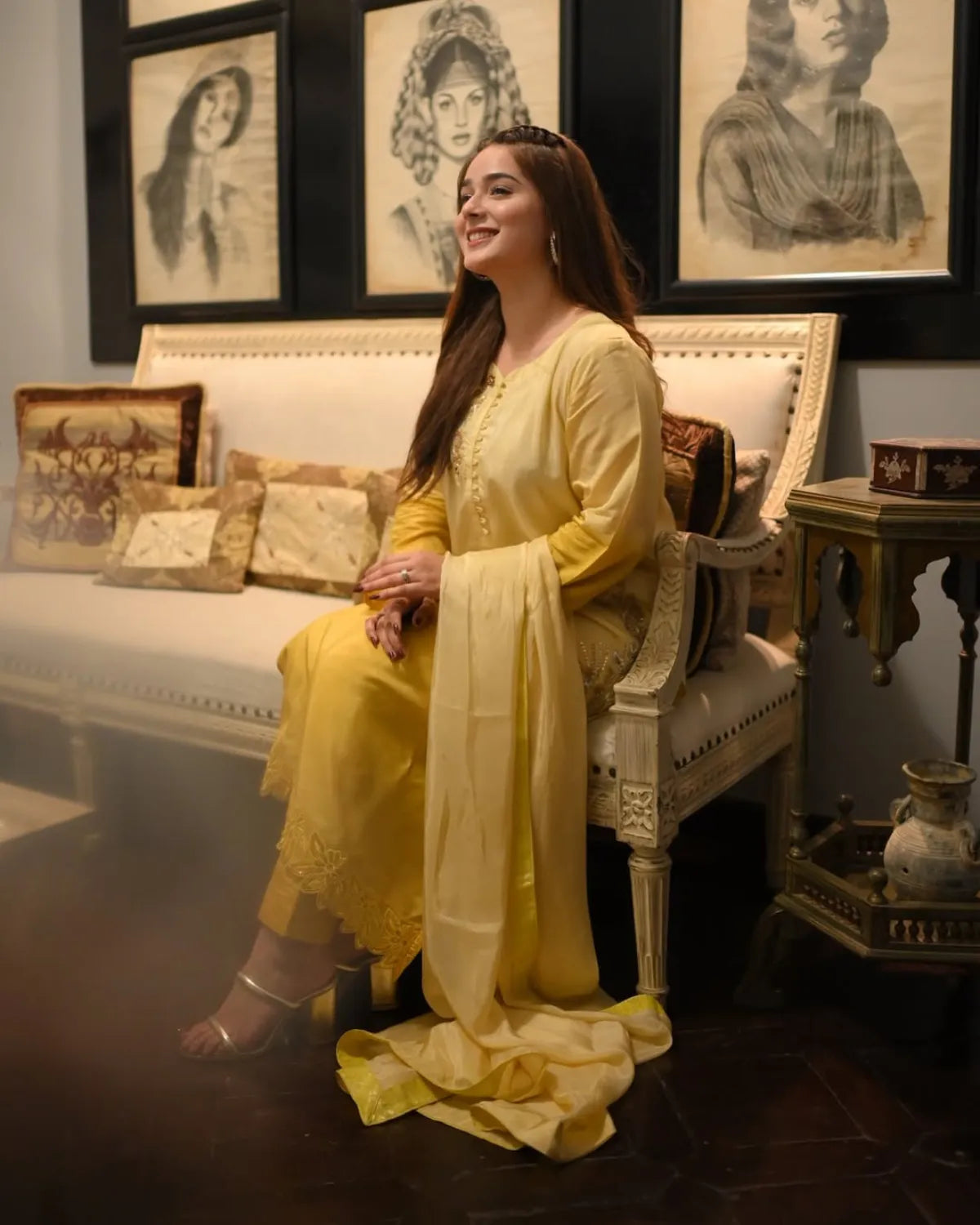 A woman wearing a traditional yellow ethnic outfit with a matching dupatta sits on an ornate white sofa in a decorated room with framed sketches on the wall.