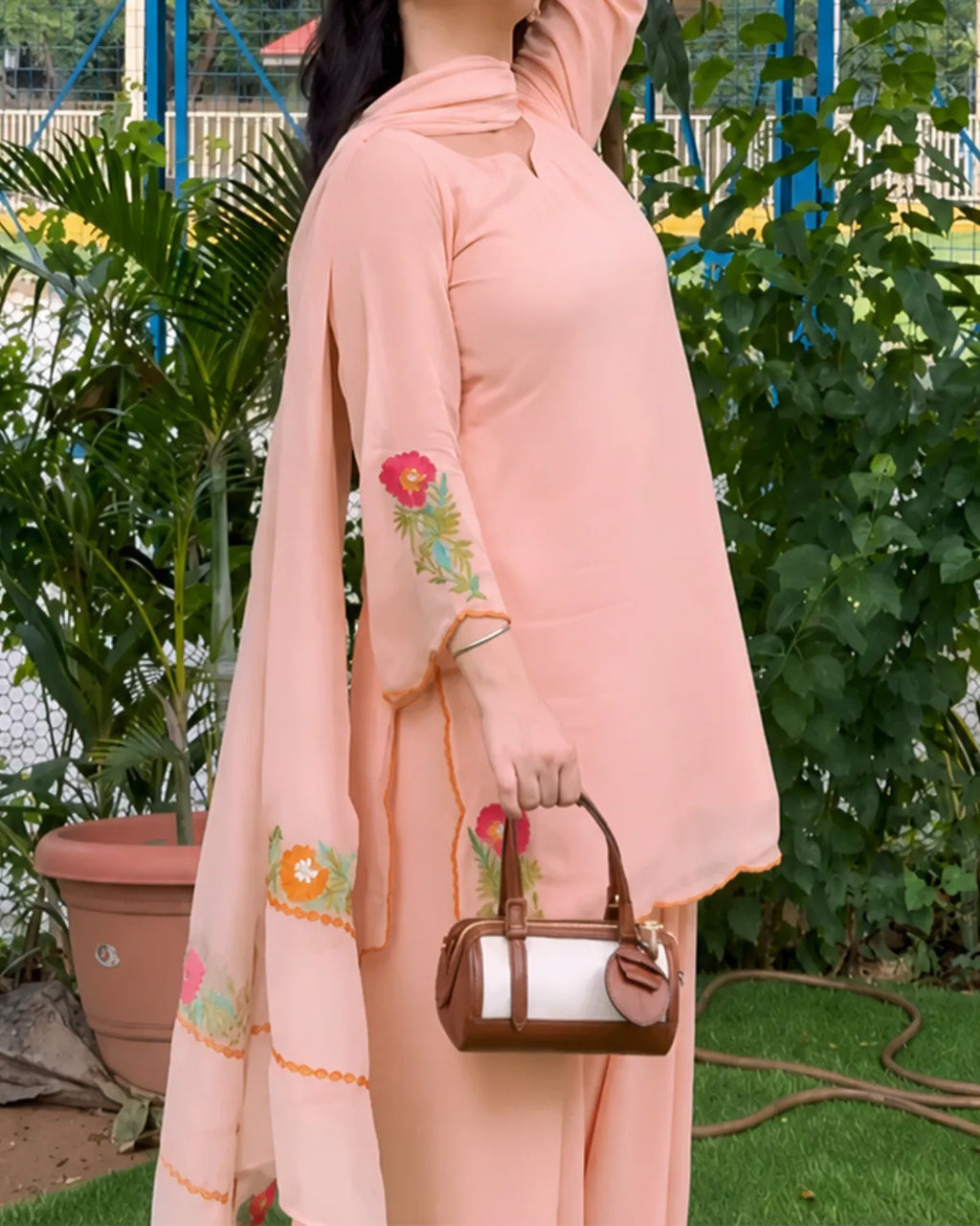 A woman wearing a peach-pink traditional outfit with floral embroidery on the sleeves and dupatta, holding a brown and white handbag in an outdoor garden setting.
