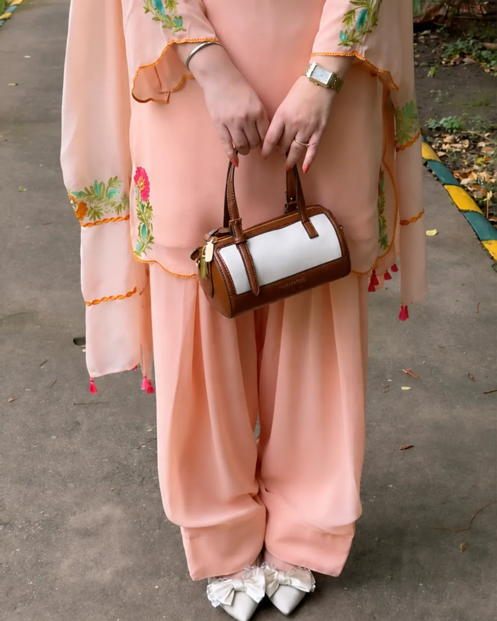 A person wearing a peach-pink traditional outfit with floral embroidery and holding a small brown and white handbag, standing outdoors on a paved path.