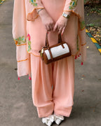A person wearing a peach-pink traditional outfit with floral embroidery and holding a small brown and white handbag, standing outdoors on a paved path.