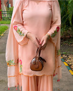 A person wearing a peach-pink traditional outfit with floral embroidery on the sleeves and hem, holding a small brown handbag outdoors on a paved path with greenery in the background.