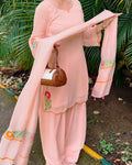 A woman wearing a light pink traditional salwar kameez suit with matching dupatta, featuring colorful floral embroidery on the sleeves and border, holding a brown handbag outdoors in front of green foliage.