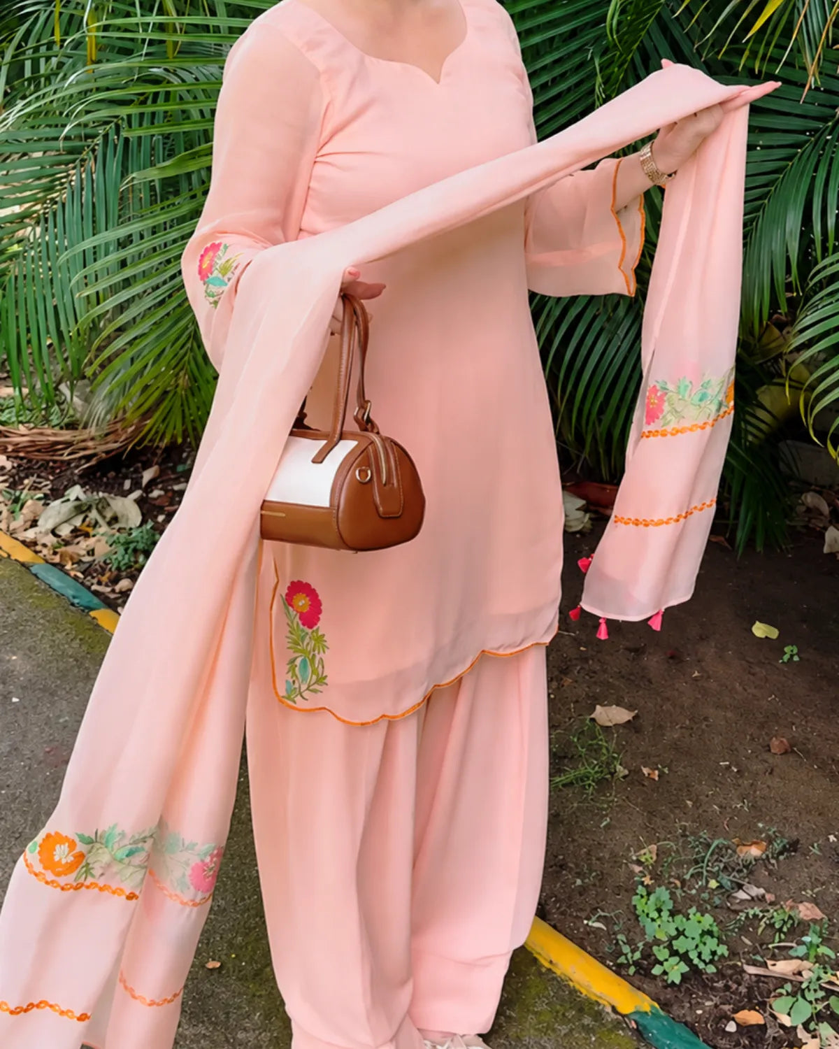 A woman wearing a light pink traditional salwar kameez suit with matching dupatta, featuring colorful floral embroidery on the sleeves and border, holding a brown handbag outdoors in front of green foliage.