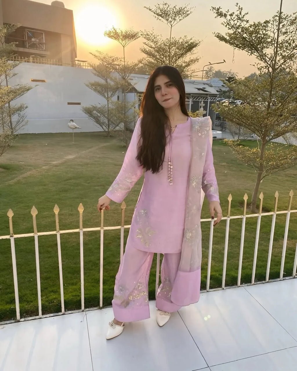 A woman wearing a light pink traditional outfit with matching pants and a sheer embroidered dupatta, standing on a balcony at sunset with a lawn and trees in the background.