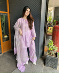 A woman wearing a light purple/lavender three-piece traditional outfit with a matching sheer dupatta, standing in front of a dark textured metal wall and doorway.