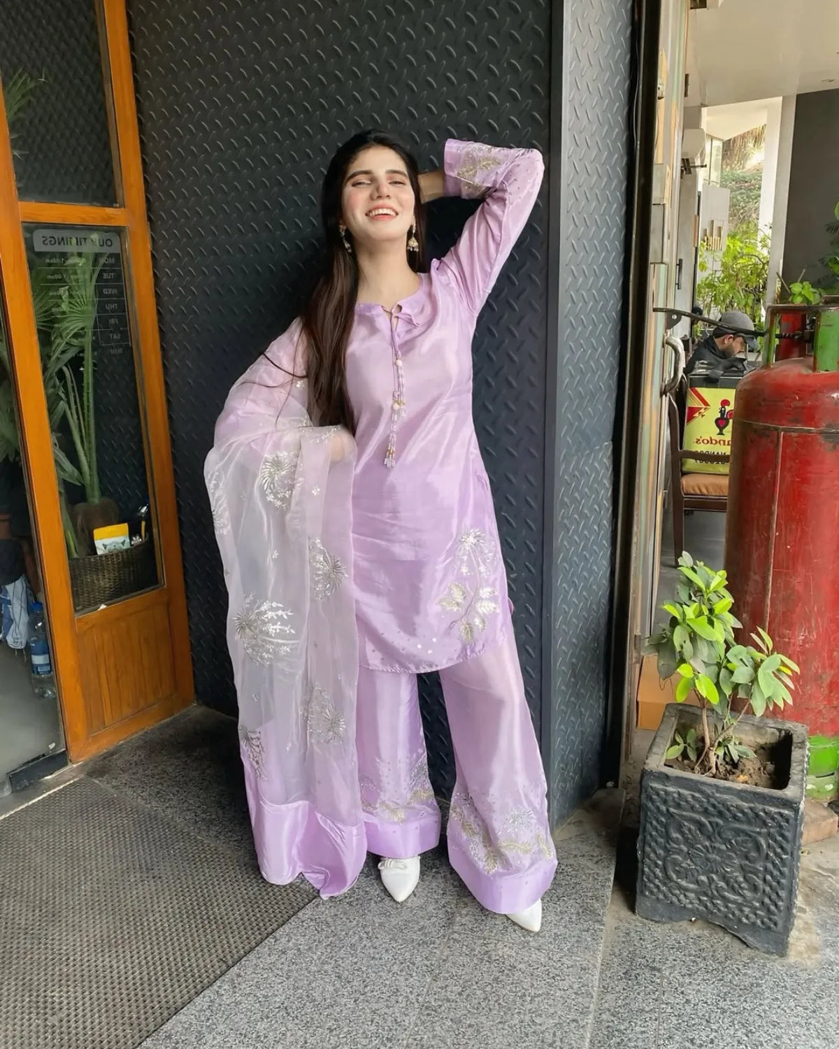 A woman wearing a light purple/lavender traditional outfit with a matching sheer dupatta and white pointed shoes, posing in front of a dark textured wall.