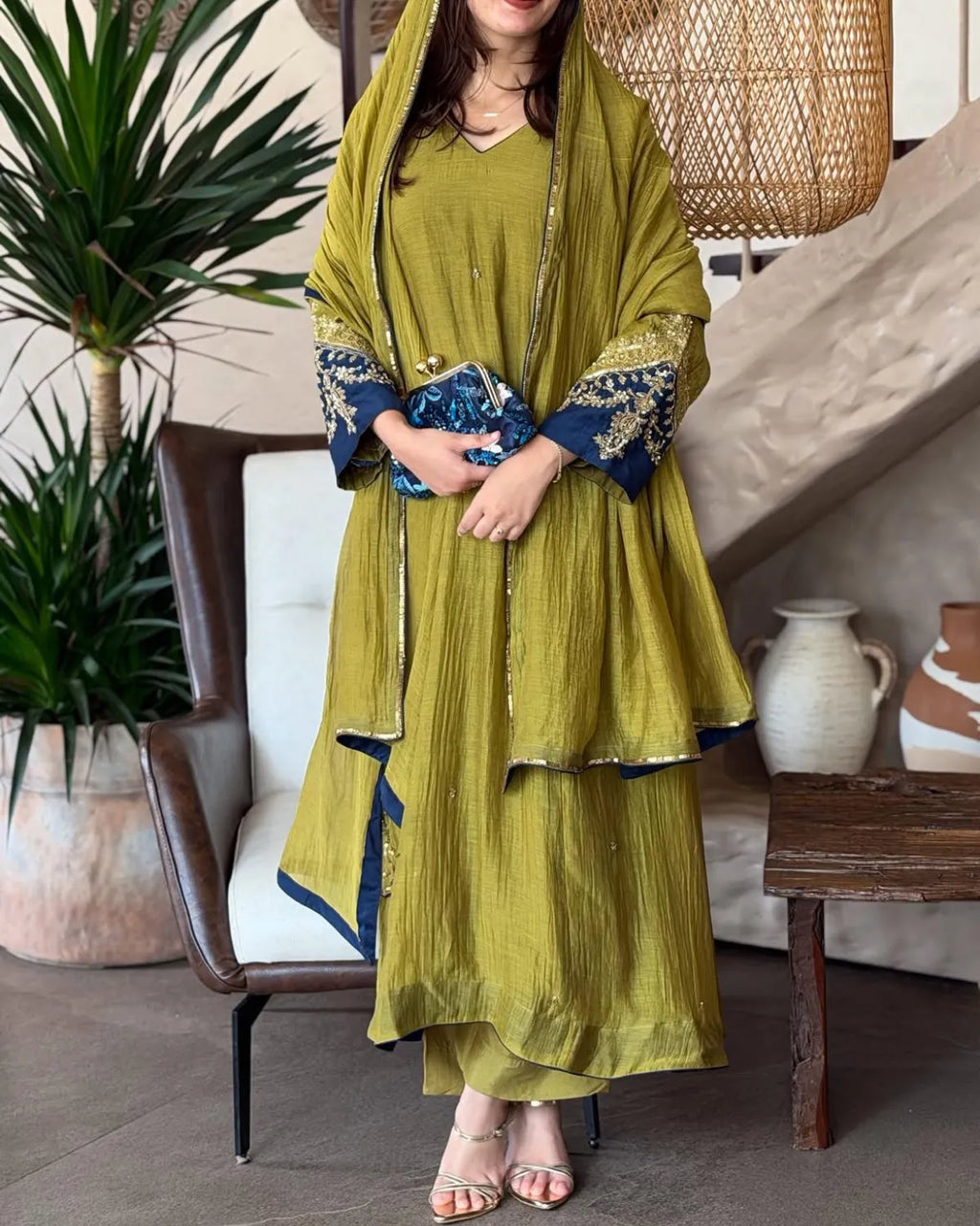 A woman models a traditional olive green salwar suit set with a matching dupatta, featuring navy blue and gold embroidered sleeve cuffs, styled in a cozy indoor setting.