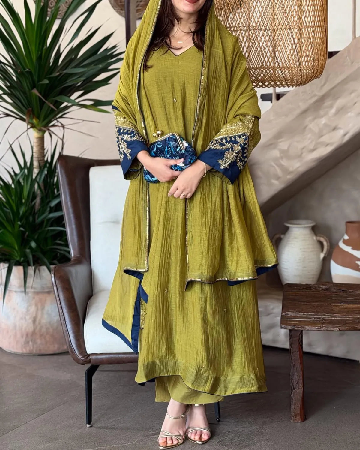 A woman models a traditional olive green salwar suit set with a matching dupatta, featuring navy blue and gold embroidered sleeve cuffs, styled in a cozy indoor setting.