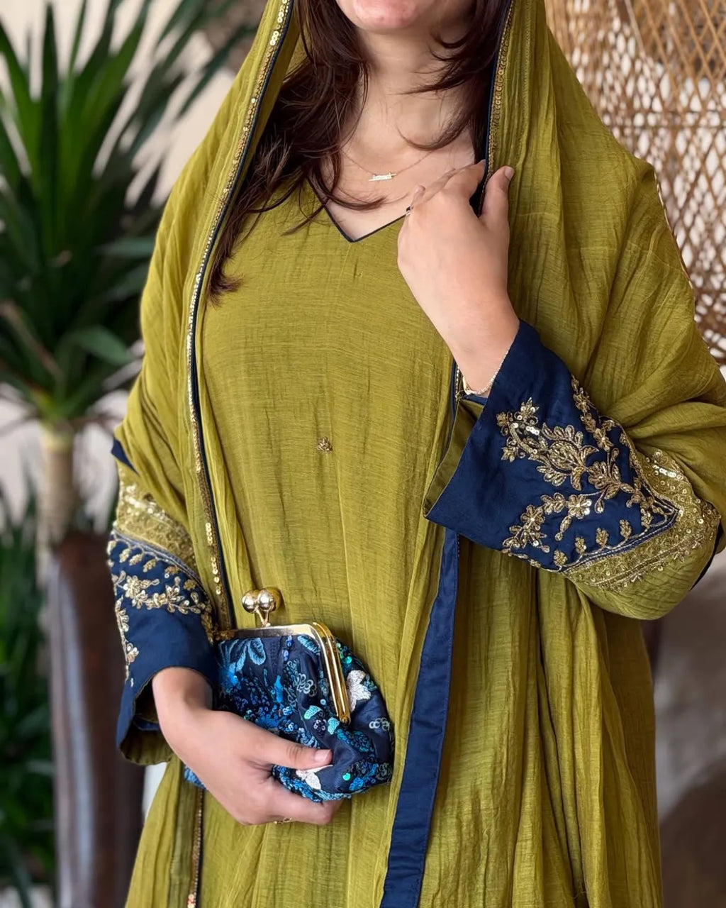 A woman wearing a mustard yellow traditional outfit with navy blue sleeves featuring gold embroidery, holding a matching navy blue clutch purse with gold hardware.