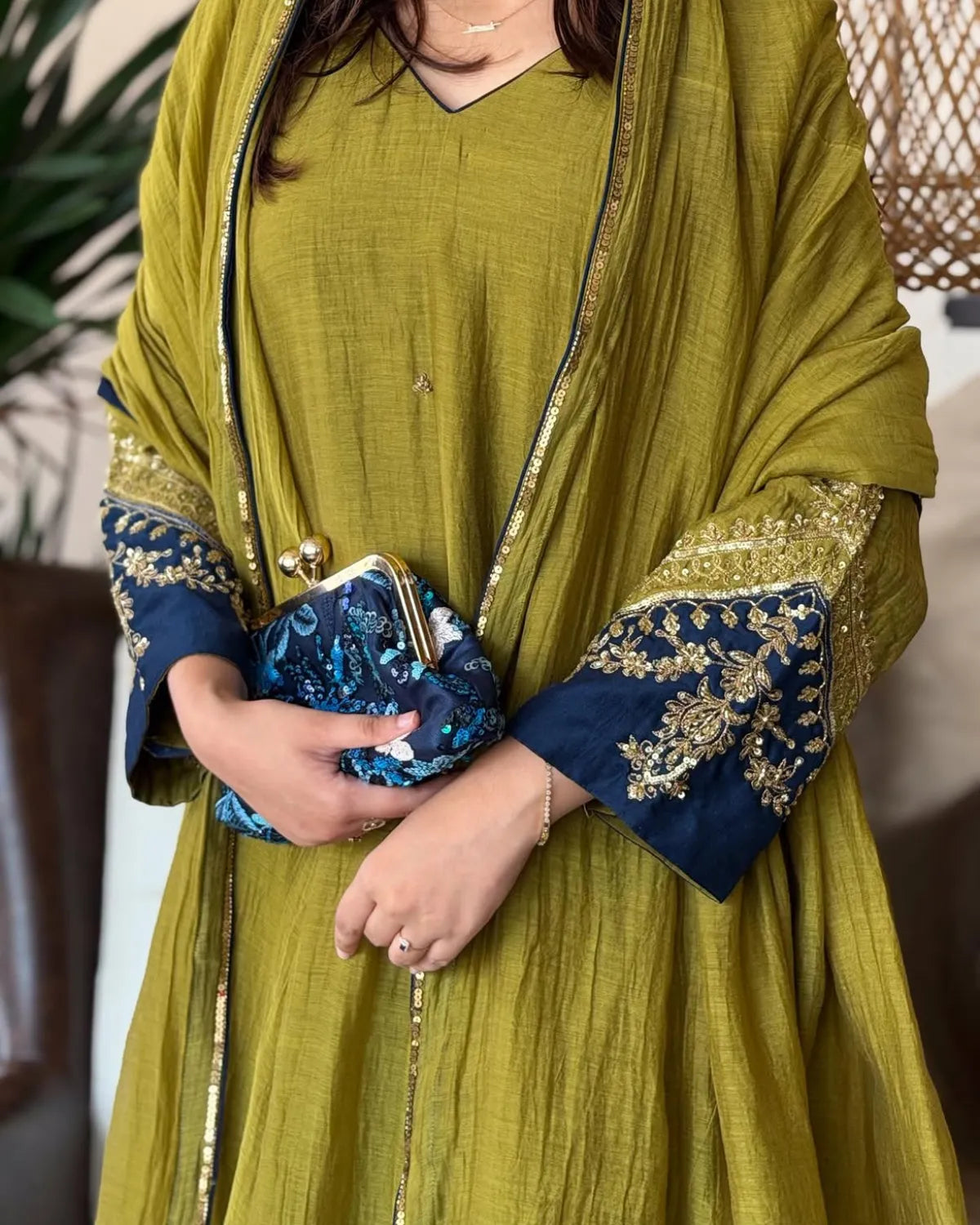 A person wearing a mustard yellow-green traditional outfit with navy blue and gold embroidered cuffs, holding a blue patterned clutch purse.