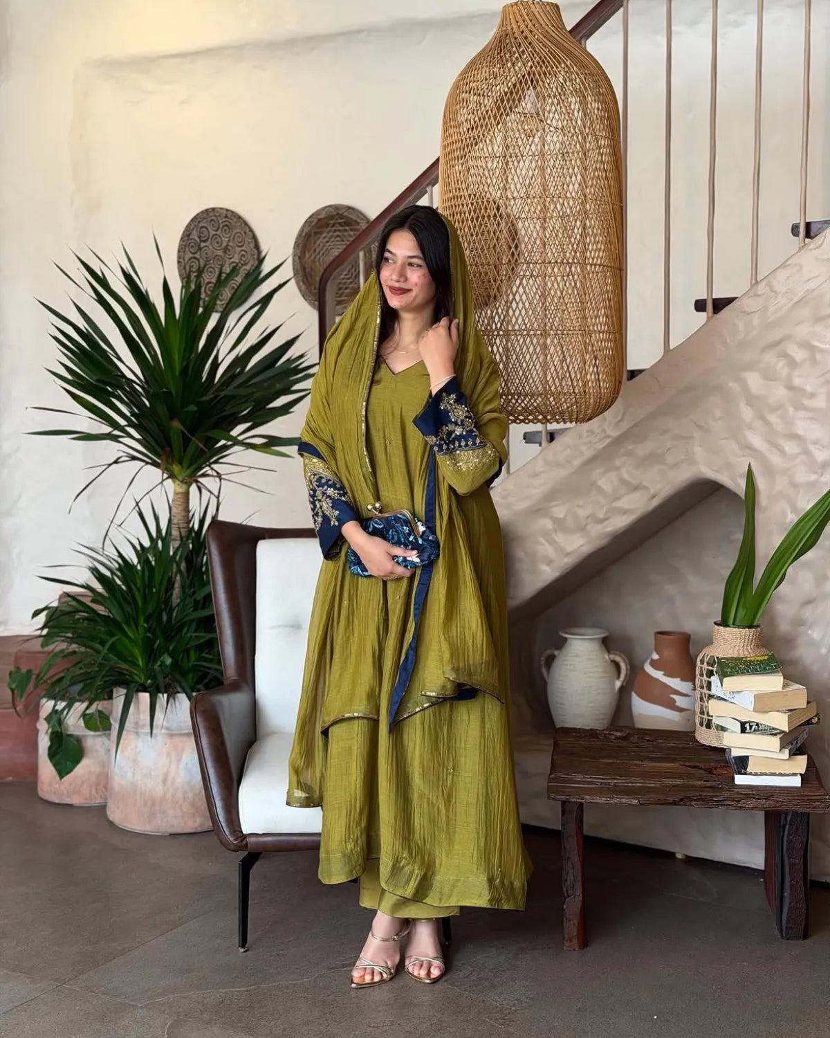 A woman models a traditional olive green salwar kameez set with a matching dupatta, featuring blue and gold embroidered sleeves, posed in a rustic, bohemian-style room with a staircase, rattan lamp, and potted plants.