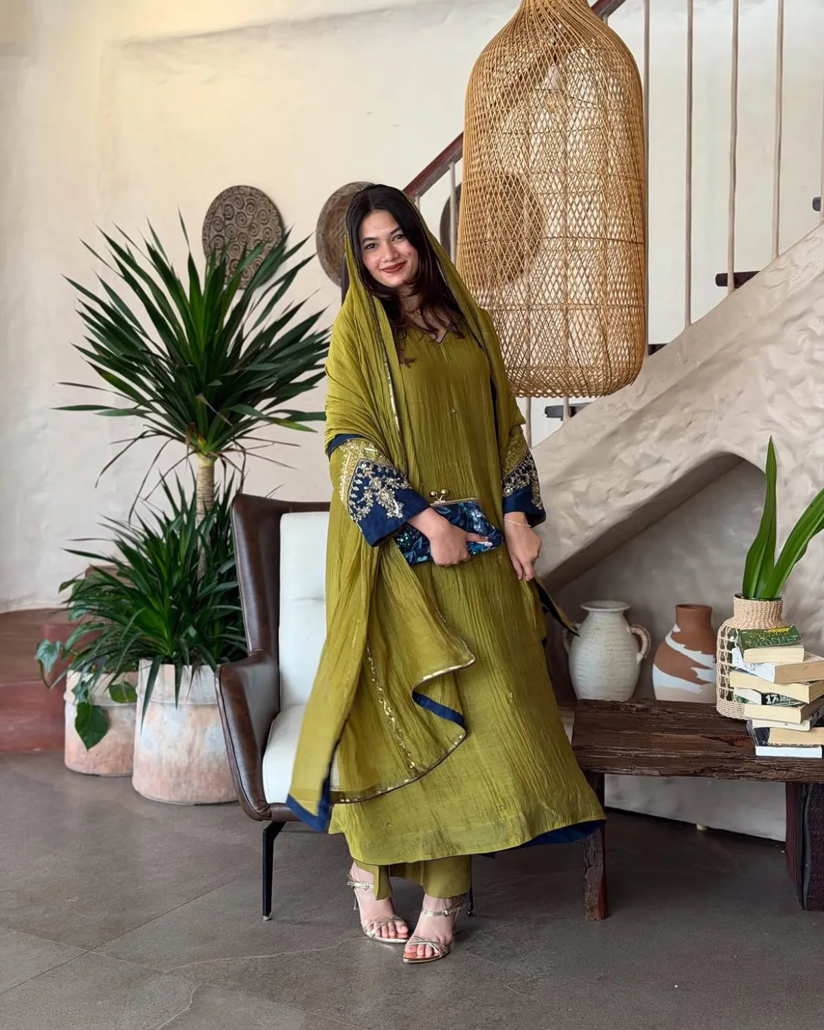 A woman models a traditional olive green three-piece salwar kameez suit with navy blue embroidered sleeves and a matching dupatta, standing in a styled indoor room with plants and a staircase.