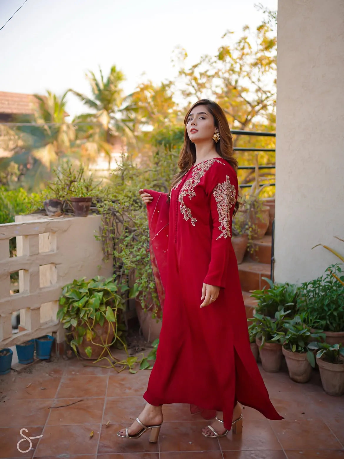 A woman wearing a long red maxi dress with gold embroidery on the chest and sleeves, standing on a tiled outdoor balcony with plants and a wall in the background.