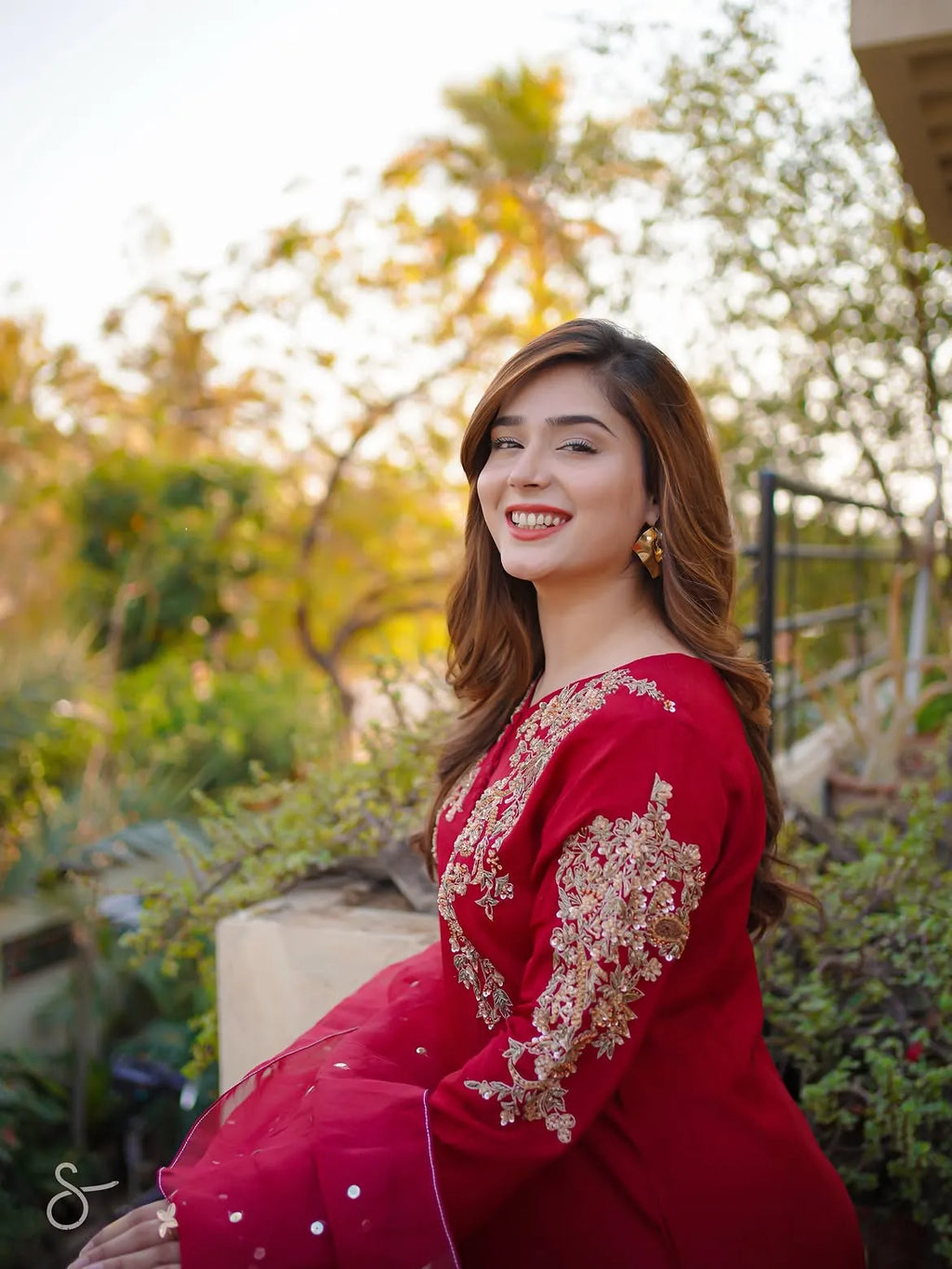 A woman wearing a red traditional outfit with gold embroidery, posing outdoors in front of a softly blurred garden background.