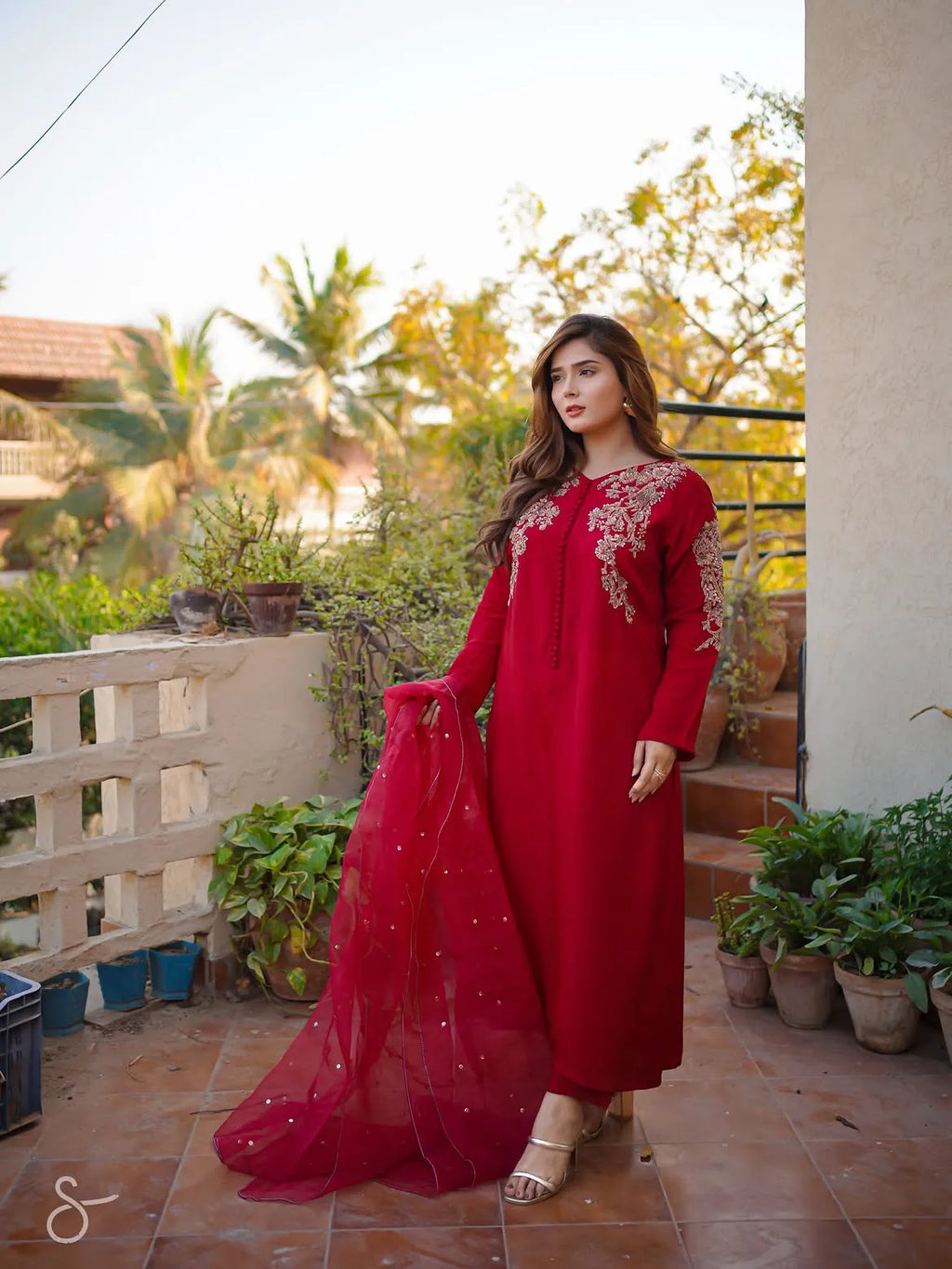 A woman models a traditional red long kurta with intricate floral embroidery on the chest and sleeves, paired with a matching sheer red dupatta, set against an outdoor patio background with potted plants.