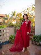 A woman models a traditional red long kurta with intricate floral embroidery on the chest and sleeves, paired with a matching sheer red dupatta, set against an outdoor patio background with potted plants.