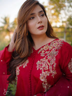 A woman wearing a red traditional embroidered outfit with gold floral detailing, photographed outdoors against a softly blurred background of trees and warm light.