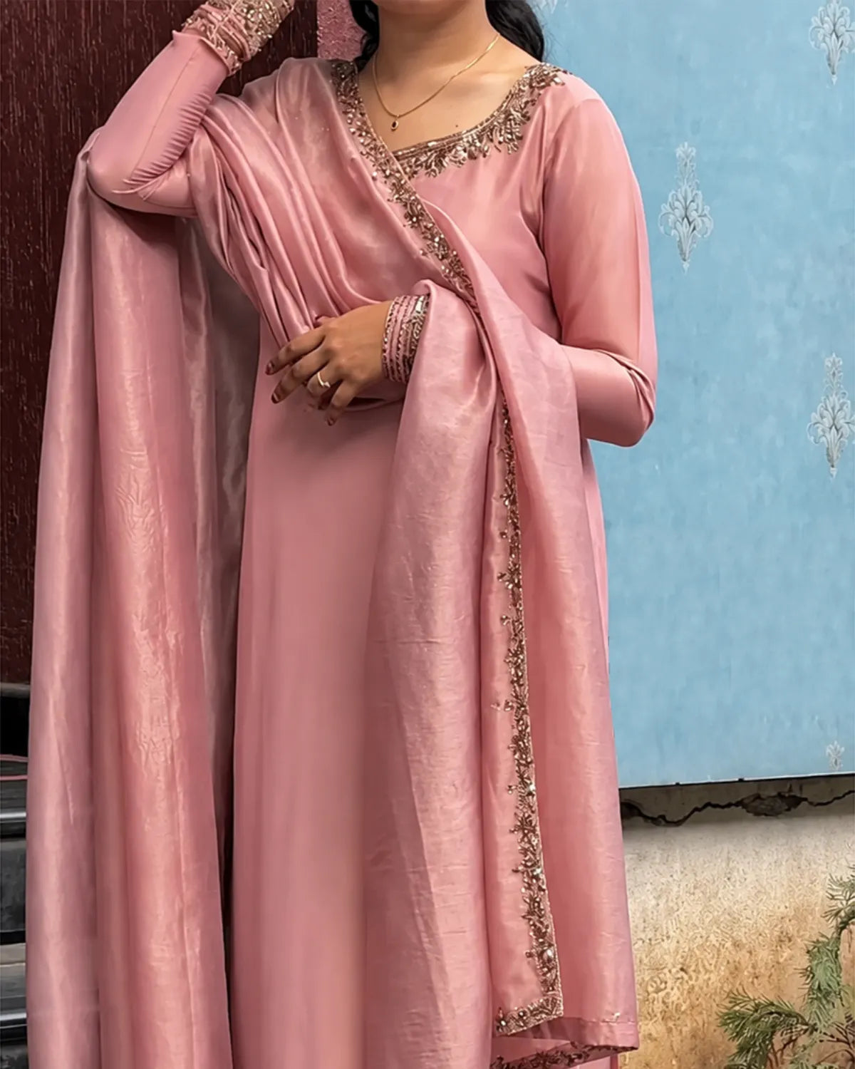 A woman wearing a dusty pink traditional outfit with a matching draped scarf, featuring gold embroidered details along the neckline and edges.