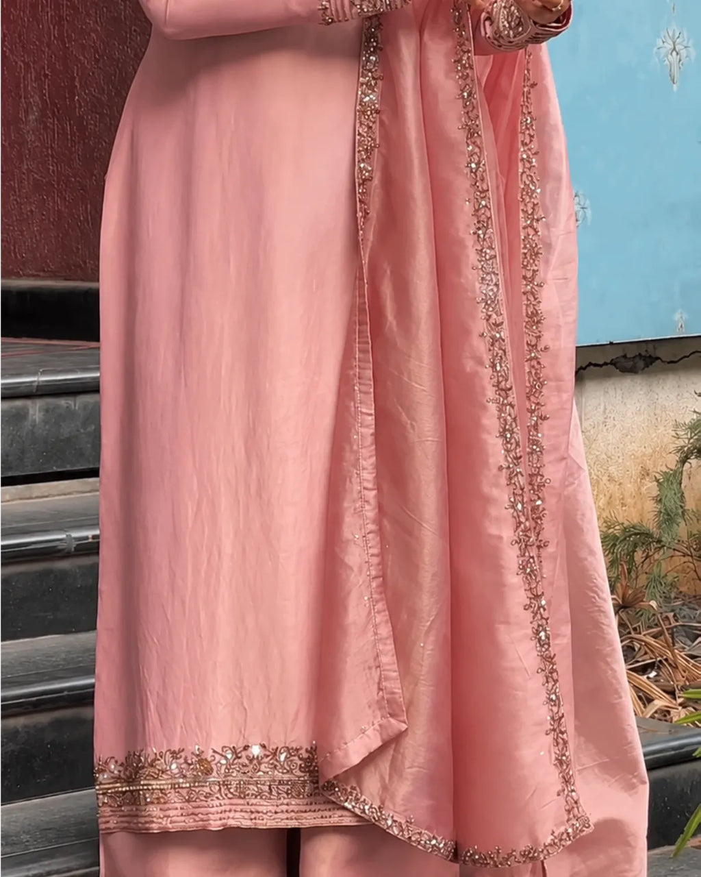 A close-up of a pink traditional outfit with matching dupatta, featuring gold and silver embroidered borders, worn by a model standing outdoors.