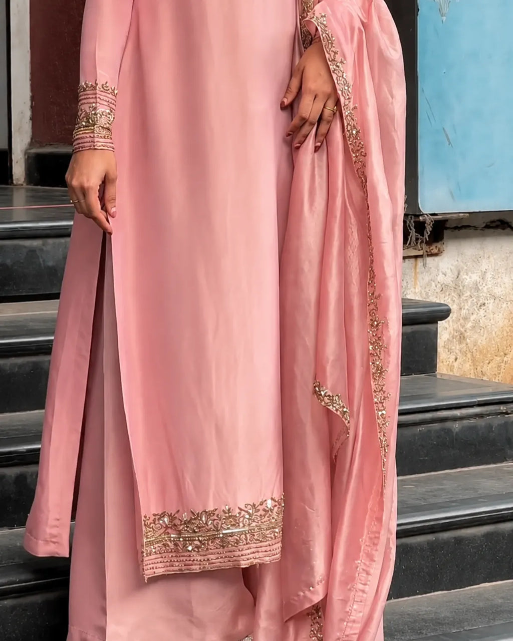A person wearing a light pink traditional outfit with gold embroidered detailing on the sleeve cuffs, hem, and draped dupatta, standing on dark outdoor steps.