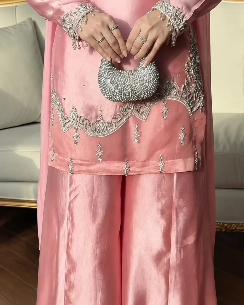A close-up of a person wearing a pink traditional outfit with silver embroidery and holding a silver beaded clutch purse in an indoor setting.