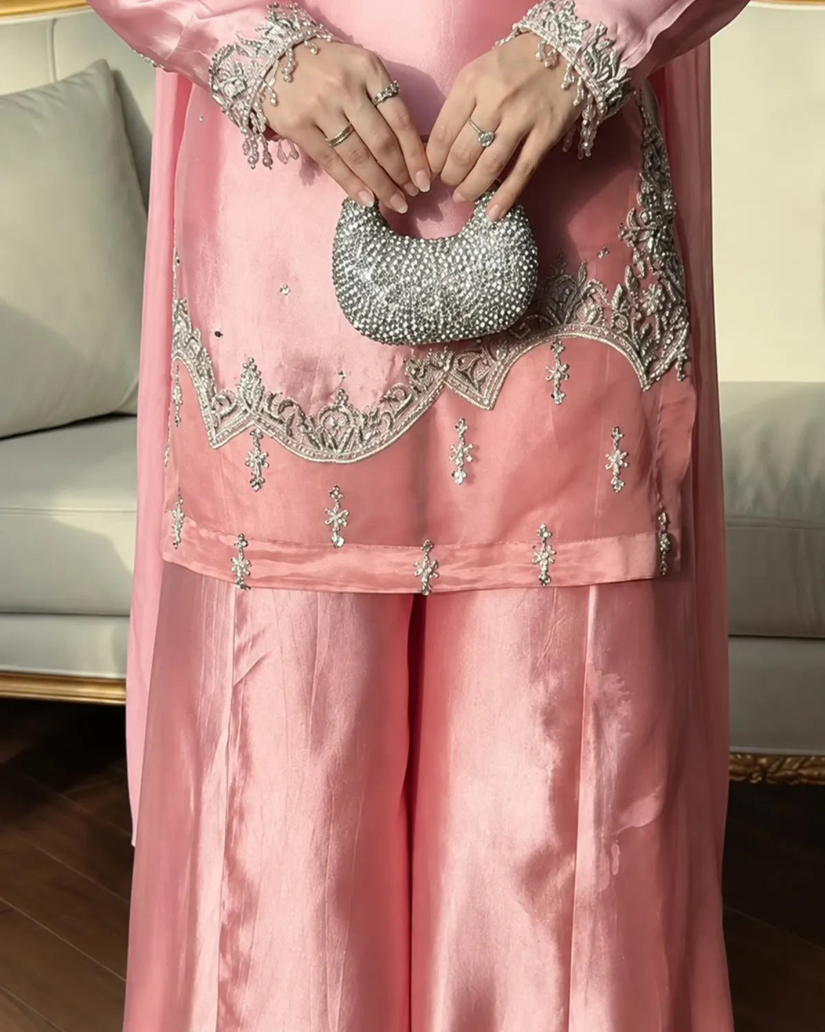 A close-up of a person wearing a pink traditional outfit with silver embroidery and holding a silver beaded clutch purse in an indoor setting.
