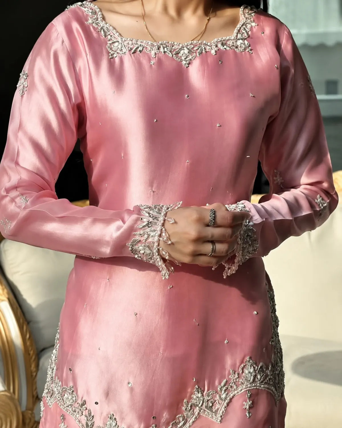 A close-up of a person wearing a pink satin traditional outfit with intricate silver and white floral embroidery and beadwork, featuring long sleeves and a square neckline.