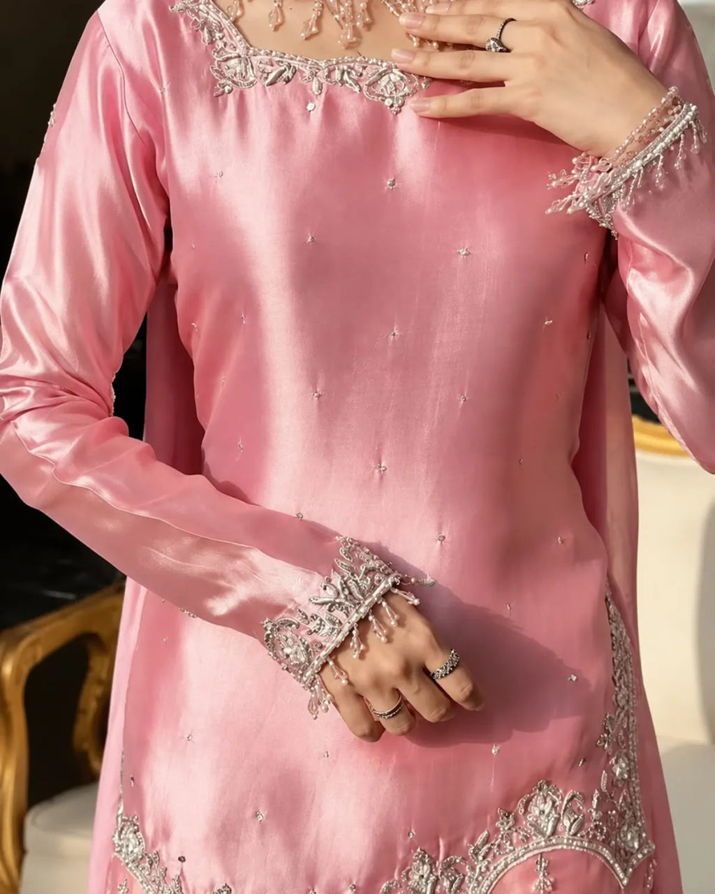 A close-up of a person wearing a light pink satin traditional outfit with intricate silver and white embroidery on the neckline, cuffs, and hem, accessorized with rings and bracelets.