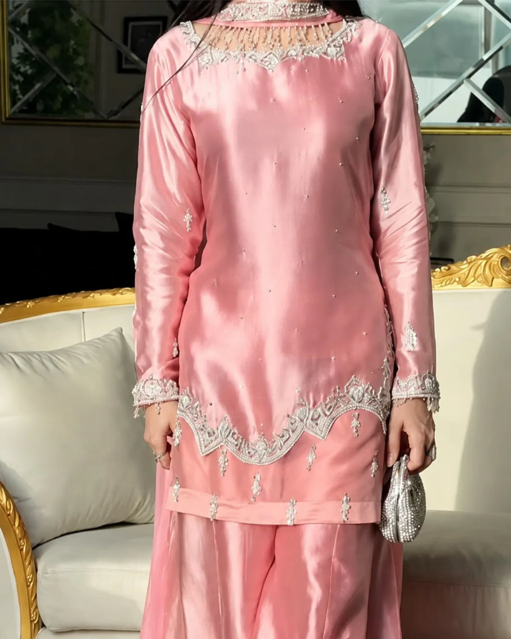 A woman wearing a pink satin three-piece traditional outfit with silver embroidery, standing in front of a cream-colored sofa with gold trim.