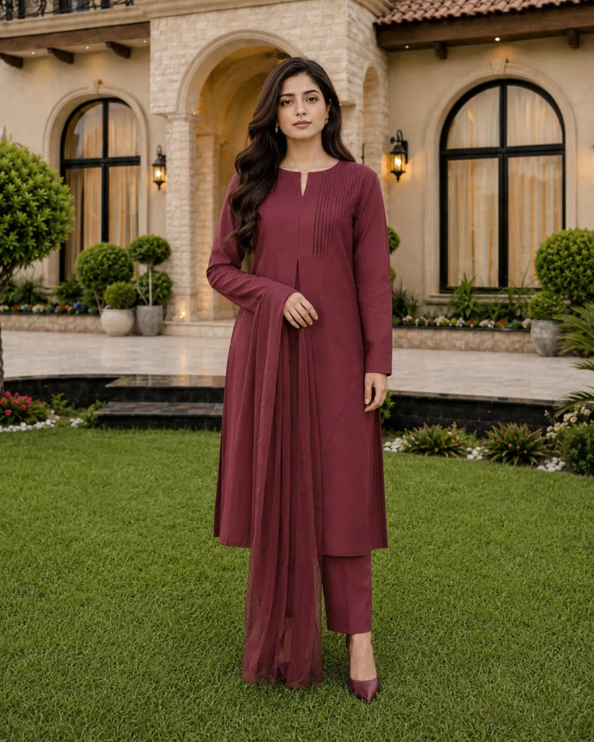 A woman stands on a green lawn in front of a large stone villa, wearing a long-sleeve maroon kurta with matching pants and pointed shoes.