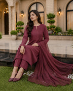 A woman wearing a long-sleeve maroon ethnic salwar kameez suit with matching dupatta sits on a stone ledge in front of a grand building with columns and green plants.