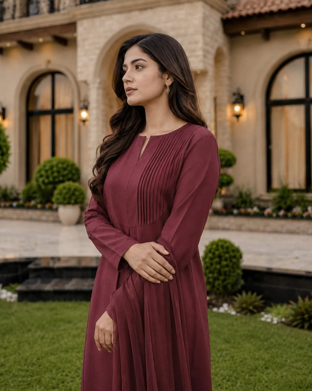 A woman wearing a long-sleeved, pleated maroon dress stands in front of a grand stone building with arched windows and manicured green hedges.