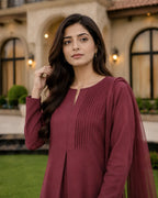 A woman wearing a long-sleeved maroon kurta with pleated front detailing and a matching sheer dupatta, standing outdoors in front of a stone building with green grass in the foreground.