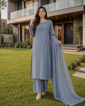 A woman stands on a green lawn in front of a modern house, wearing a light blue traditional outfit with a matching draped scarf.