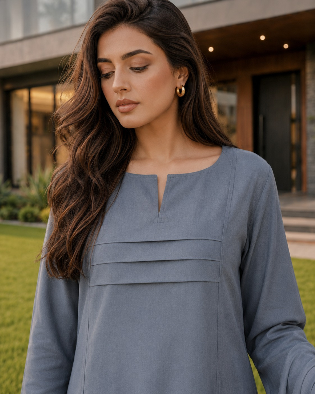 A woman wearing a slate blue long-sleeve tunic top with a split neckline, standing outdoors in front of a modern building with green grass in the background.