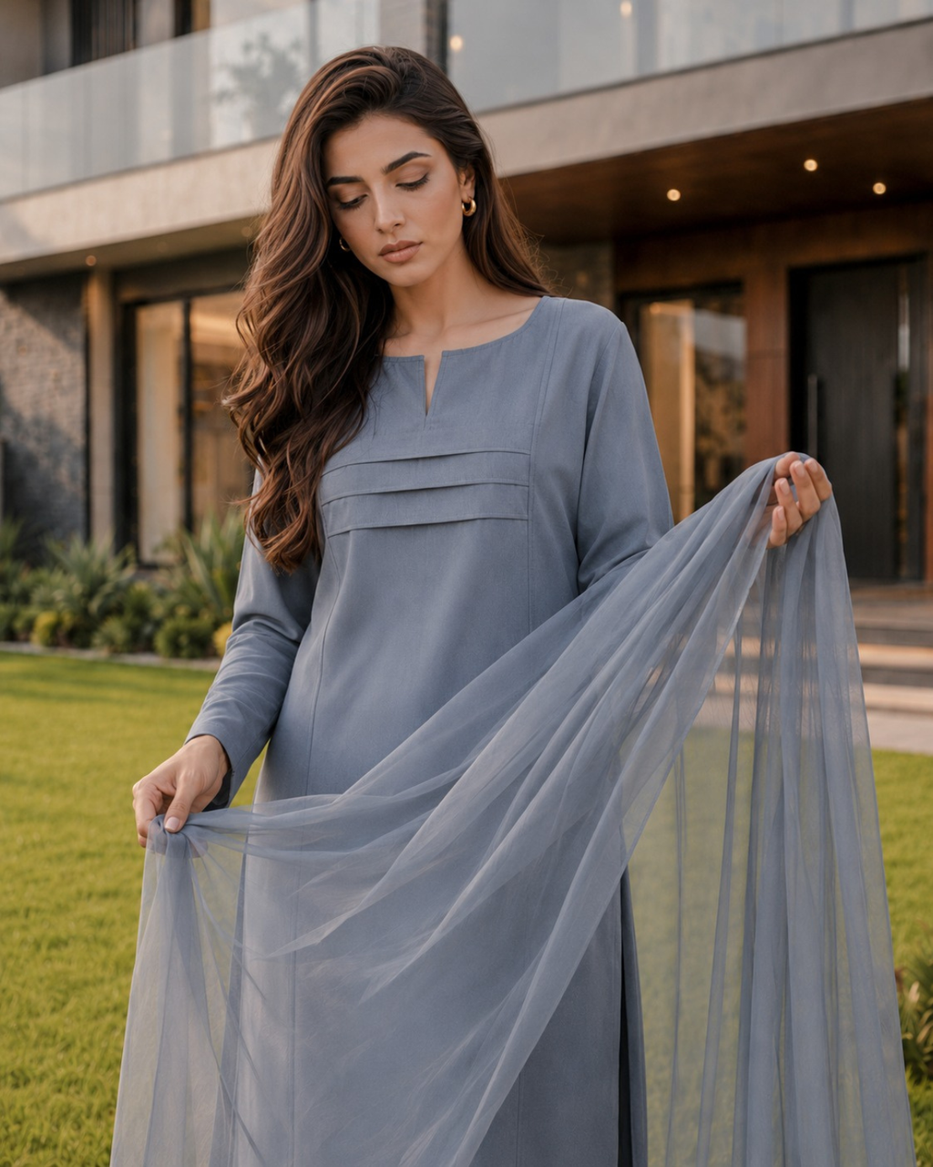 A woman wearing a long-sleeve dusty blue-grey tunic dress with pleated detailing at the chest, holding up a matching sheer dupatta in an outdoor garden setting.