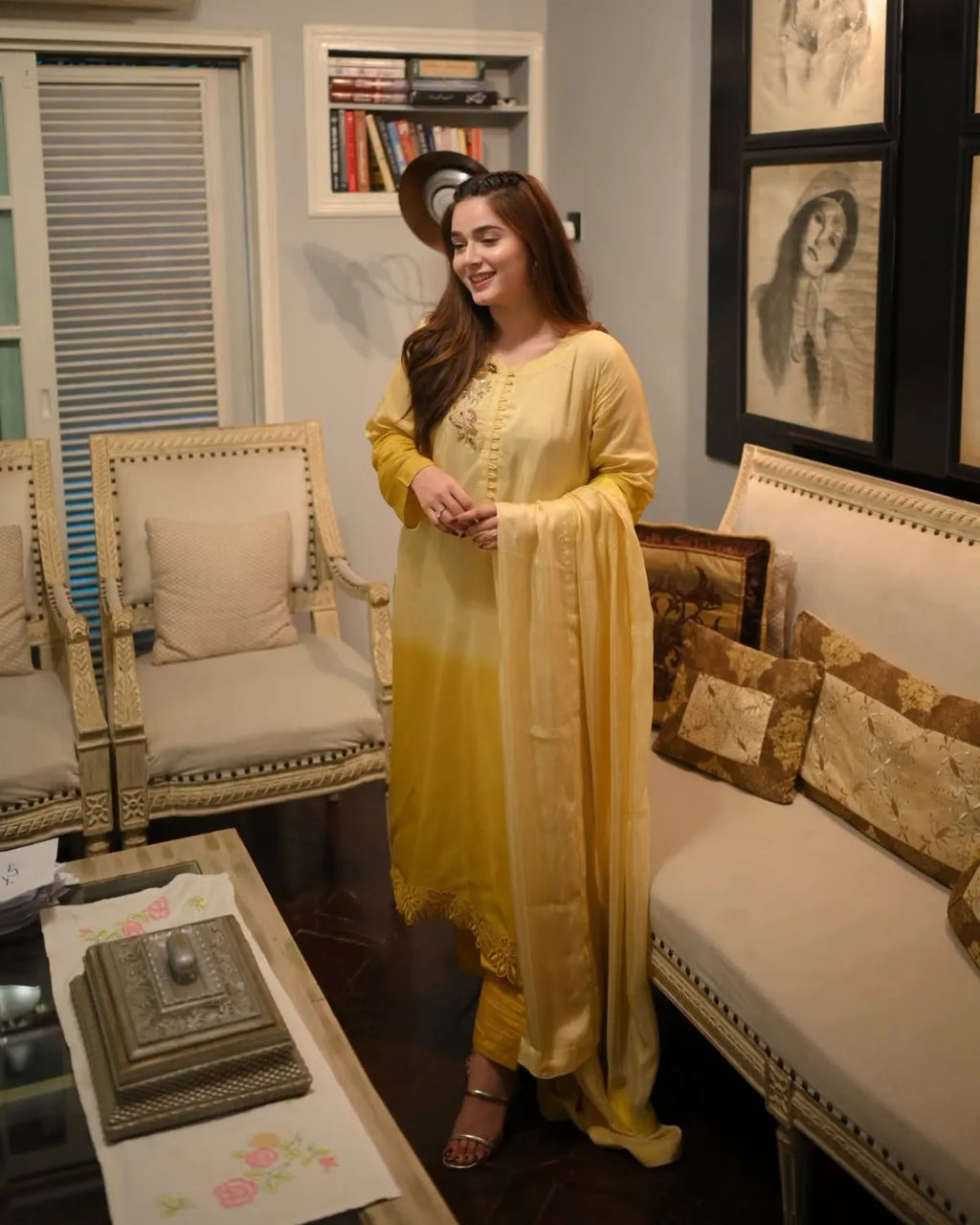 A woman wearing a yellow traditional South Asian outfit with a matching dupatta stands in a furnished living room with cream-colored armchairs, a sofa, framed wall art, and a coffee table.