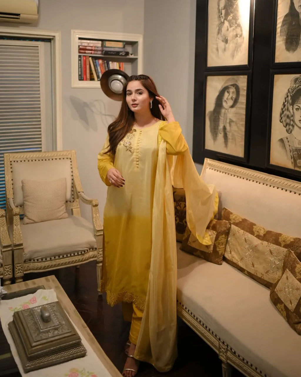 A woman stands in a furnished living room wearing a yellow traditional outfit with matching pants and a sheer draped overlay. The room includes a sofa with patterned cushions, a chair, framed wall art, and a small table in the foreground.