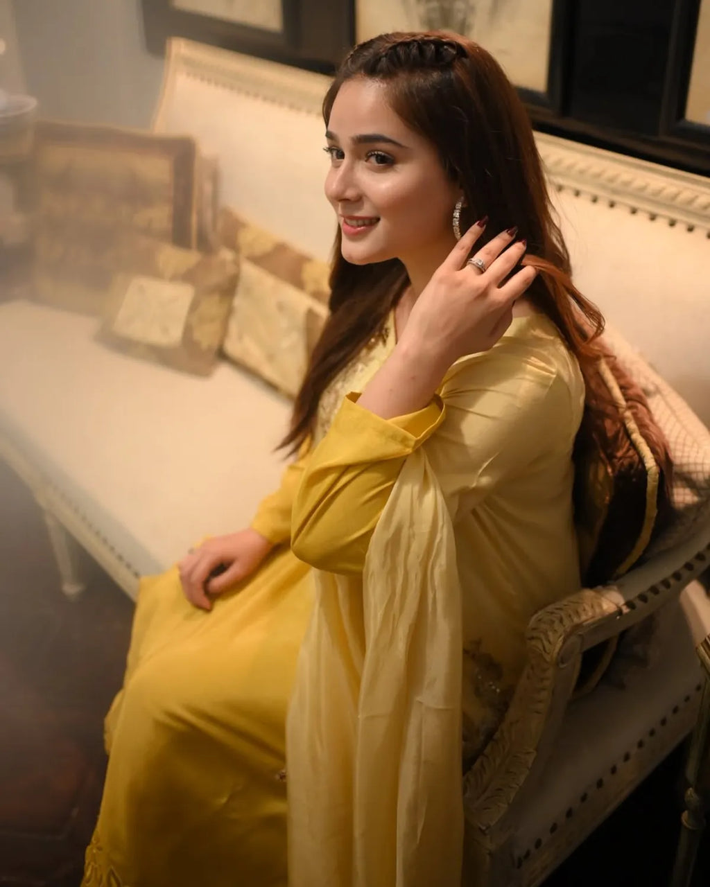 A woman wearing a yellow traditional outfit sits on a chair in an indoor setting with a sofa and pillows in the background.
