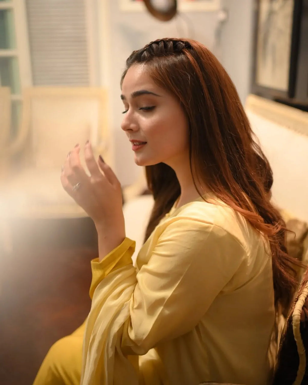 A woman with long brown hair wearing a yellow traditional outfit, captured in a warm indoor setting with soft lighting.