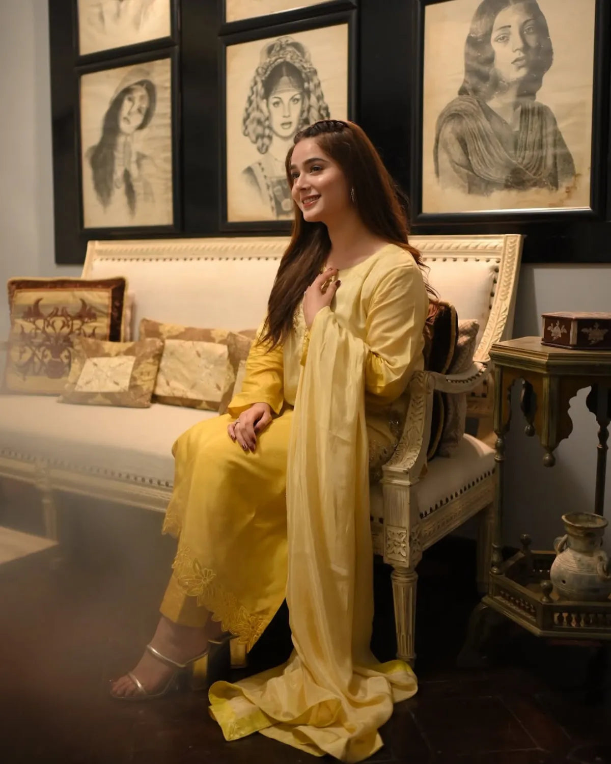 A woman wearing a flowing yellow traditional outfit sits on an ornate cream-colored sofa in a decorated room with framed black-and-white portraits on the wall.