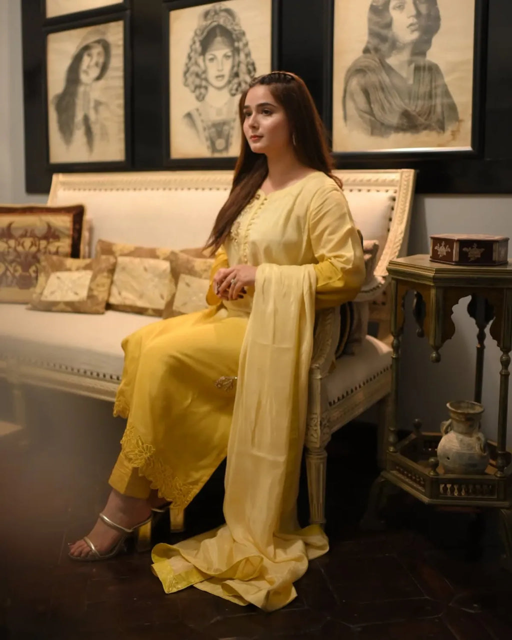 A woman wearing a traditional yellow salwar kameez suit with a matching dupatta sits on an ornate cream-colored sofa in a decorated room with framed sketches on the wall.