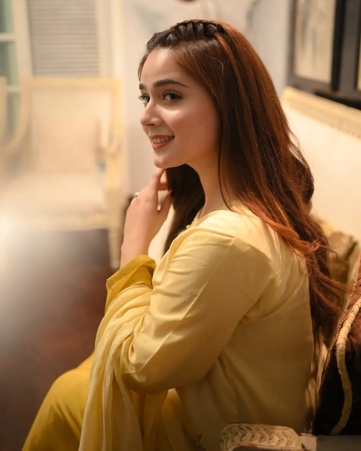 A woman with long brown hair wearing a light yellow traditional outfit, sitting in a softly lit indoor setting.